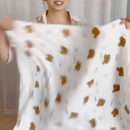 Une personne tient et plie un Lange Bebe | TyHelloBear™ blanc présentant un motif de visage d'ours brun et un petit texte, avec des rideaux légers et une partie de leur visage et de la partie supérieure de leur corps en arrière-plan.