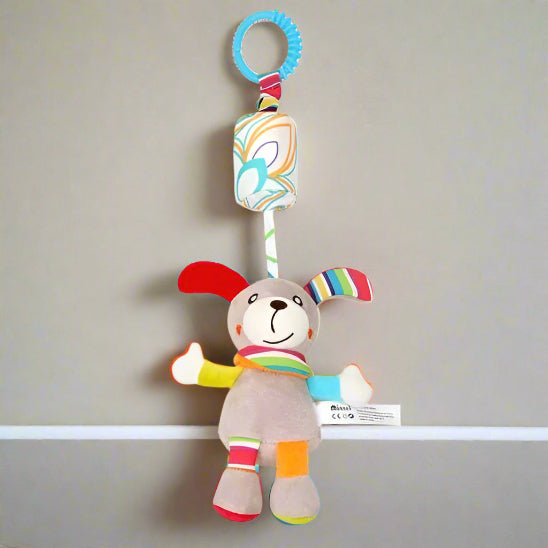Le Jouet - Hochet Bébé est un chien en peluche coloré avec des oreilles tombantes, des patchs lumineux pour l'éveil sensoriel, et est attaché à une pince à motifs par une sangle en tissu, présentée suspendue sur un fond gris clair uni.