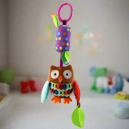 Un Jouet - Hochet Bébé en peluche colorée, avec des ailes à motifs et un haut à pois, est suspendu à un anneau rose. L'arrière-plan légèrement flou montre une chambre d'enfant joyeuse, idéale pour l'éveil sensoriel.