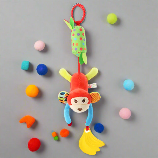 Un Jouet - Hochet Bébé vibrant avec un singe en peluche et une banane est suspendu à une boucle rouge, entouré de balles colorées sur un fond gris - idéal pour l'éveil sensoriel de bébé.