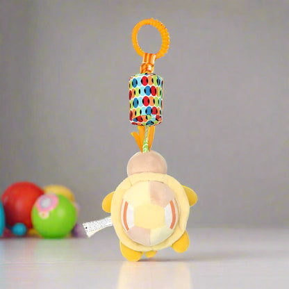 Le Jouet - Hochet Bébé est une tortue jaune en peluche avec une carapace hexagonale, suspendue à un anneau orange et à une attache colorée, parfaite pour l'éveil sensoriel. Des boules floues sont visibles en arrière-plan sur une surface blanche.
