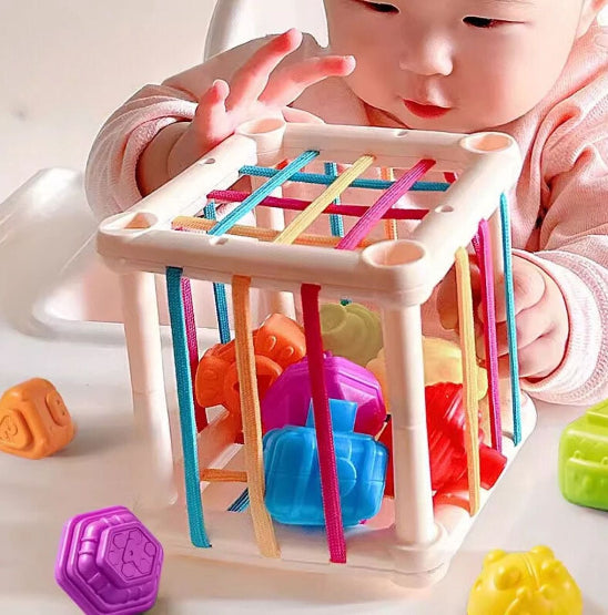 Un bébé découvre les Jeux d'éveils - cubes éducatifs, un cube sensoriel en plastique blanc avec bandes élastiques multicolores et blocs géométriques, idéal pour stimuler le développement et l'éveil sur une surface blanche.
