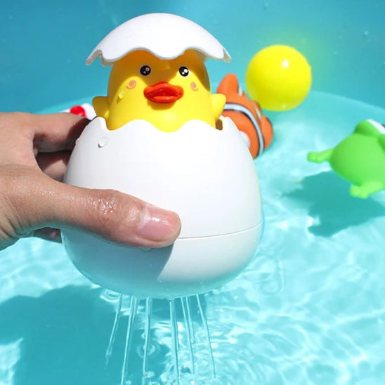 Une main tient le canard en caoutchouc jaune « Jouet de bain » à l'intérieur d'une coquille d'œuf blanche, immergé dans l'eau, tandis qu'un jet d'eau s'abat sur lui. Des jouets flottants colorés, comme une grenouille et un poisson-clown, sont visibles en arrière-plan.