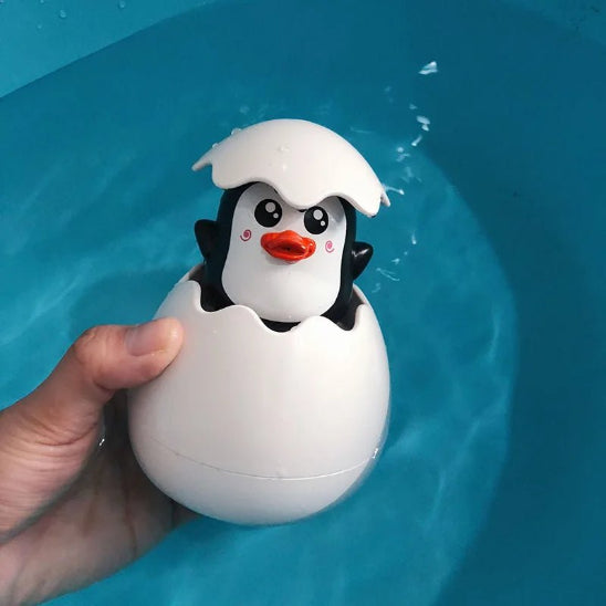 Une main tient le Jouet de bain - Jet d'eau pour Enfants, un jouet de bain en forme de pingouin émergeant d'une coquille d'œuf blanche, avec un visage souriant et un bec orange - idéal pour rendre l'heure du bain amusante et ludique au-dessus de l'eau bleue.