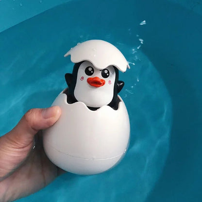 Une main tient le Jouet de bain - Jet d'eau pour Enfants, un jouet de bain en forme de pingouin émergeant d'une coquille d'œuf blanche, avec un visage souriant et un bec orange - idéal pour rendre l'heure du bain amusante et ludique au-dessus de l'eau bleue.
