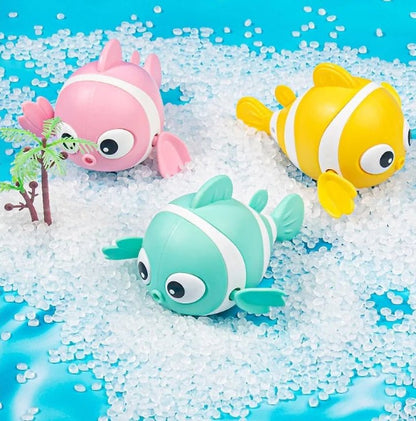 Trois jouets de bain - des poissons nageurs roses, jaunes et verts menthe avec des rayures blanches reposent sur des granulés blancs à côté d'un petit palmier en plastique, sur un fond bleu qui évoque le plaisir du bain de bébé.