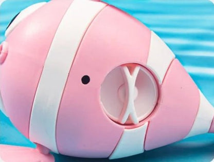 Gros plan du Jouet de bain – poisson nageur, un jouet en forme de poisson rayé rose et blanc, doté d'une ouverture circulaire ornée d'un X blanc. Sur fond bleu rayé. Idéal pour baigner bébé !.