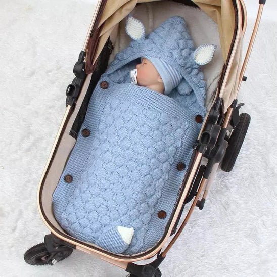 Un bébé est allongé dans une poussette, confortablement installé dans le Nid d'Ange Bébé Hiver - Couverture Câline et Douillette bleue à capuche avec de petites oreilles. Le bébé porte un bonnet bleu et a une tétine. La poussette est posée sur une surface blanche.