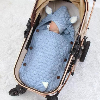 Un bébé est allongé dans une poussette, confortablement installé dans le Nid d'Ange Bébé Hiver - Couverture Câline et Douillette bleue à capuche avec de petites oreilles. Le bébé porte un bonnet bleu et a une tétine. La poussette est posée sur une surface blanche.