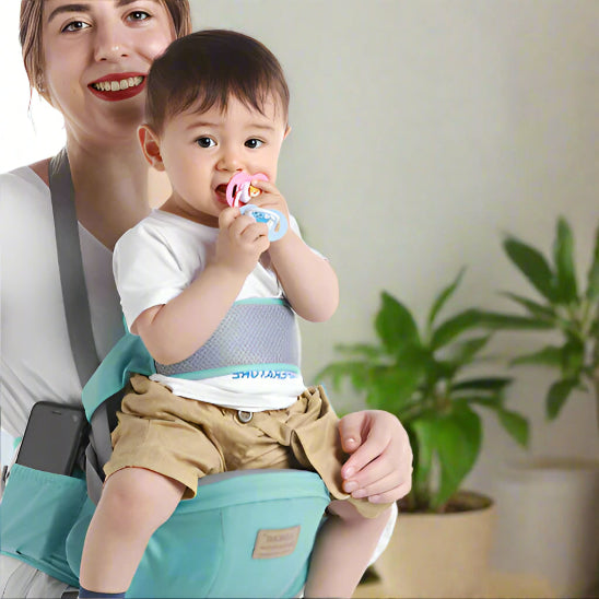 Une femme souriante porte son bébé dans le Porte-Bébé ergonomique | WaistHolder™. Le bébé, vêtu d'une chemise blanche et d'un short kaki, tient une tétine. Des plantes vertes en pot sont visibles en arrière-plan.