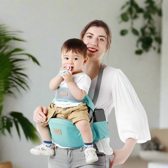 Une femme sourit en tenant un bébé dans le porte-bébé Porte Bébé | WaistHolder™, qui offre un confort ergonomique. Le bébé, vêtu d'une chemise blanche et d'un short kaki, tient une tétine. Des plantes vertes et un fond clair complètent la scène.