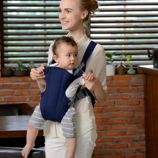Une femme sourit à l'intérieur, portant un bébé dans le porte-bébé ergonomique Porte Bébé | Face & Dos. Le bébé porte une tenue grise rayée. Derrière eux, un mur de briques, des stores en bois et des plantes en pot sur une étagère.