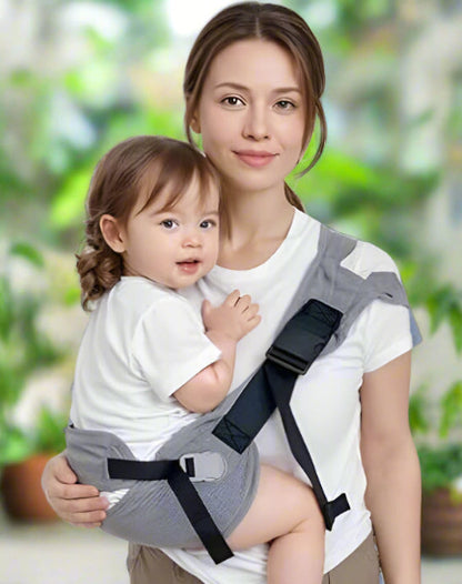 Une femme en chemise blanche porte un tout-petit HandsFree sur sa hanche dans le Porte Bébé - HandsFree gris, tous deux face à la caméra avec un fond vert doux, idéal pour les parents actifs.