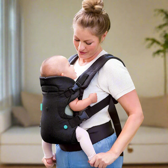 Une femme porte son bébé face à elle dans le Porte Bébé physio | FlipAdvanced™, vêtue d'un t-shirt blanc et d'un pantalon bleu. Un canapé et une plante dans le salon lumineux apportent un confort optimal à la mère et à l'enfant.
