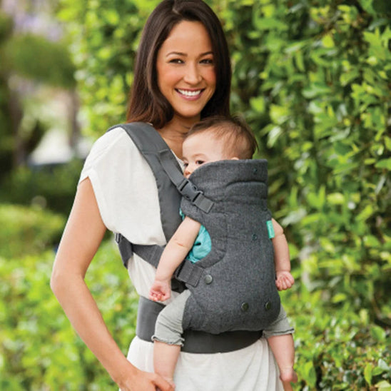 Une femme en chemise blanche se tient dehors, souriante, portant son bébé dans un Porte-Bébé physio gris | FlipAdvanced™. Un feuillage vert luxuriant forme un décor serein.