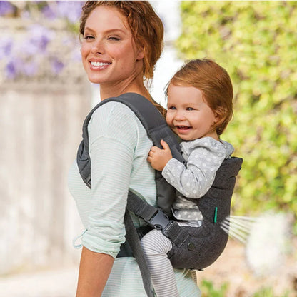 Une femme porte un tout-petit souriant sur son dos, vêtue d'un Porte-Bébé physio gris | FlipAdvanced™. Tous deux sont dehors, au milieu de la verdure ; elle porte un t-shirt rayé et l'enfant une tenue grise à pois pour un confort optimal.