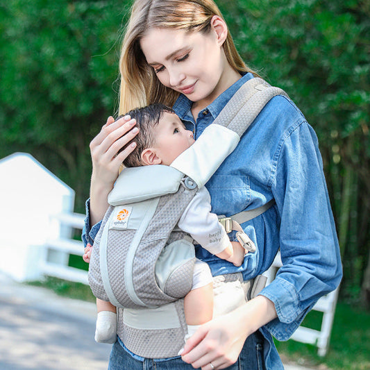 Une femme portant une chemise en jean bleu porte son bébé à l'extérieur dans un Porte Bébé physiologique | AirMesh™ gris, touchant doucement la tête du bébé alors qu'elle regarde vers le bas, entourée de verdure.
