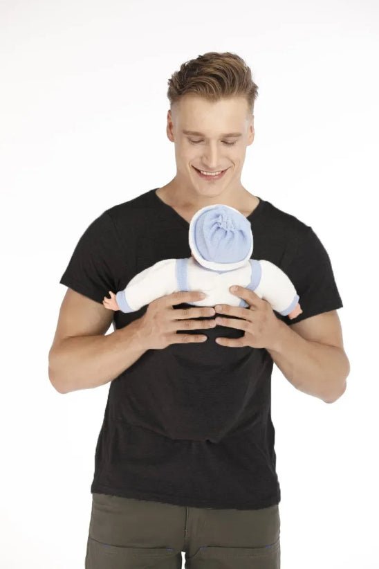 Un homme en t-shirt noir tient un bébé vêtu de blanc avec des touches de bleu et coiffé d'un chapeau bleu, tous deux tournés vers lui. Souriant au bébé, il lui présente le T-shirt Porte Bébé, une solution idéale et confortable pour les parents actifs. Le fond est blanc uni.