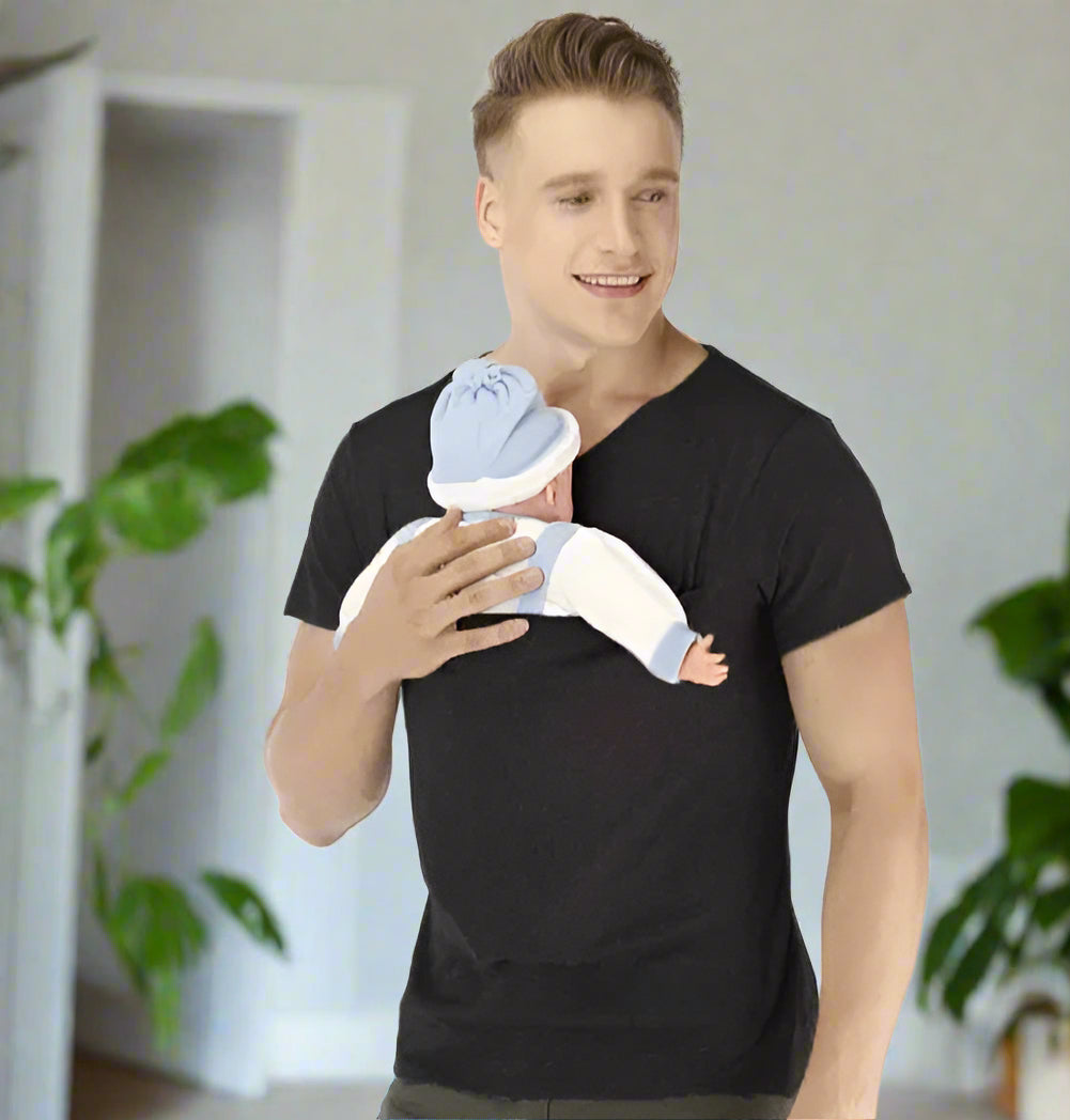Un homme portant un T-shirt « Porte-Bébé » tient un bébé vêtu de bleu et de blanc contre sa poitrine, réconfortant les parents actifs. Il sourit doucement, regardant vers la gauche, avec un arrière-plan blanc uni derrière eux.