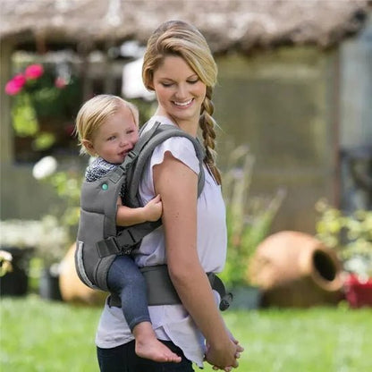 Une femme avec une tresse sur le côté porte un jeune enfant sur son dos dans le Porte Bébé | Unisexe. Elles sont dehors, dans un jardin entouré de plantes vertes et de pots en terre cuite, l'air détendues.