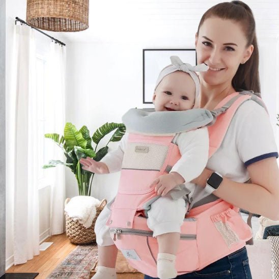 Une femme sourit dans un salon lumineux, portant son bébé dans un Porte-Bébé | WaistStool™ rose. Le bébé, confortablement installé, porte un bandeau, tandis que des plantes et une lampe en osier créent une atmosphère chaleureuse et sécurisante.
