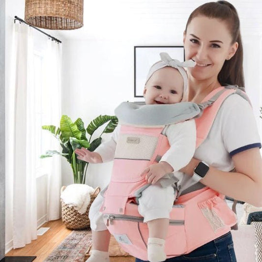Une femme sourit dans un salon lumineux, portant son bébé dans un Porte-Bébé | WaistStool™ rose. Le bébé, confortablement installé, porte un bandeau, tandis que des plantes et une lampe en osier créent une atmosphère chaleureuse et sécurisante.