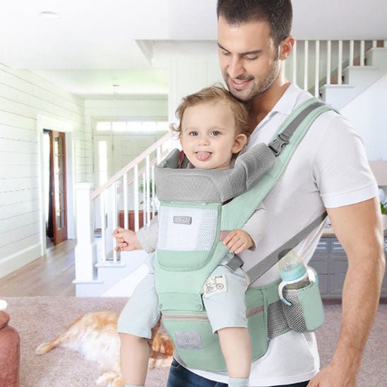 Un homme dans un salon porte un tout-petit souriant dans un Porte Bebe | WaistStool™ vert menthe avec un biberon dans la poche latérale, tandis qu'un chien est allongé sur le sol près de l'escalier.