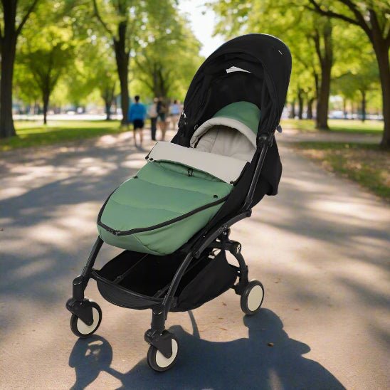 Une poussette chancelière aux motifs verts et noirs, avec un coussin blanc à protection thermique, est posée sur une allée bordée d'arbres. Deux personnes marchent à l'arrière-plan sous la canopée verdoyante.