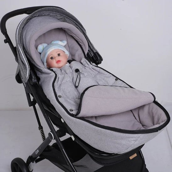 Un bébé vêtu d'une combinaison grise avec un chapeau bleu et blanc est allongé dans la Poussette Chancelière, qui offre une protection thermique, sur un fond blanc uni.