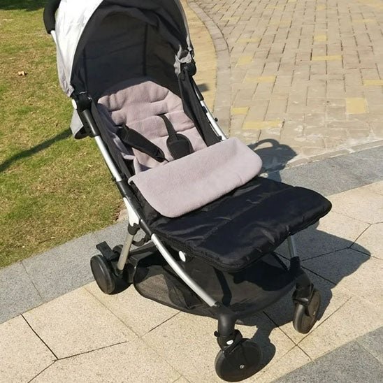 La Poussette Chancelière | CosyToes™ noire et grise est posée sur un chemin pavé au bord de l'herbe, la lumière du soleil illuminant son coussin d'assise et son harnais de sécurité. Elle protège bébé et lui assure un confort hivernal lors des journées fraîches.