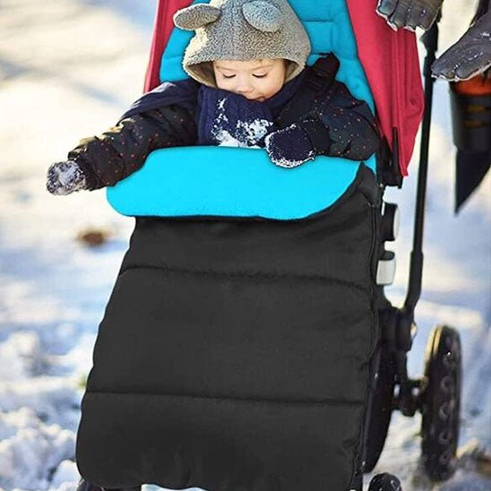 Un bébé, vêtu chaudement et coiffé d'un bonnet, est assis dans une poussette à la housse bleue et noire, équipée de la Poussette Chancelière CosyToes™ pour un confort hivernal optimal. La neige recouvre le sol, tandis qu'un adulte se tient à proximité et tient la poignée.