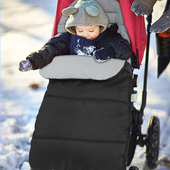 Un bébé, habillé pour l'hiver, est assis dans une poussette CosyToes™, confortablement installé dans son cocon douillet. Sa main gantée touche la neige, tandis que celle d'un adulte, gantée, tient la poignée au milieu d'un paysage enneigé.