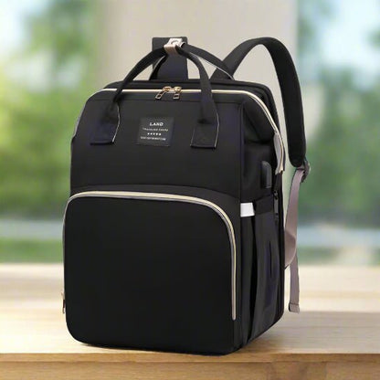 Le Sac a dos a Langer | Lit de Sortie bébé, noir avec des fermetures éclair et des poignées beiges, est présenté sur une surface claire. Il comporte des poches frontales et latérales ainsi qu'une étiquette sur le devant. L'arrière-plan légèrement flou met en valeur ce sac à langer pratique.