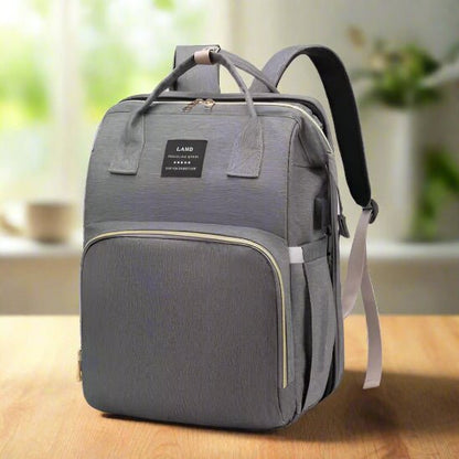 Un Sac a dos a Langer | Lit de Sortie bébé en gris avec des bretelles noires et une poche frontale zippée repose sur une table en bois d'intérieur. Une petite étiquette noire orne le sac, photographié dans une lumière douce avec un arrière-plan flou.