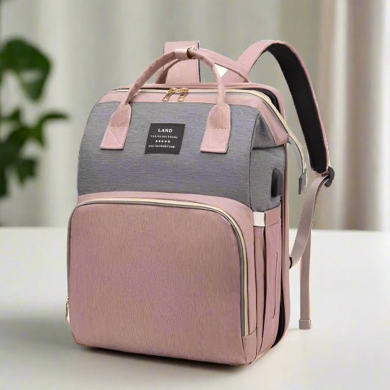 Le Sac a dos a Langer | Lit de Sortie bébé est un sac à dos à langer bicolore avec un dessus gris, un fond et des bretelles rose pâle, des poignées supérieures, une poche frontale et une étiquette plaquée noire, présentée sur une surface blanche.