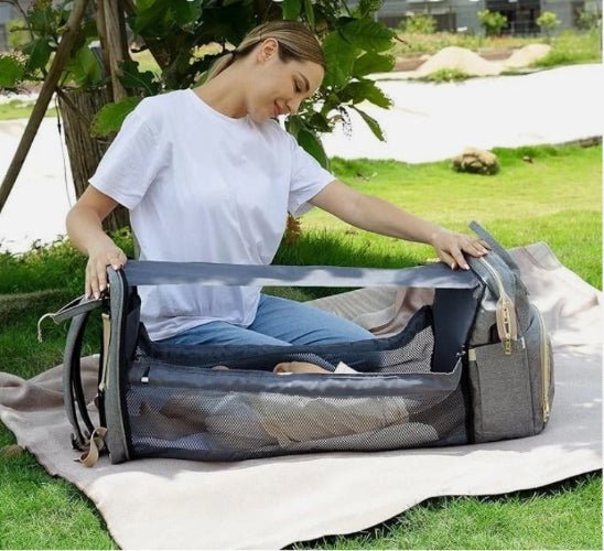 Une femme en t-shirt blanc et en jeans installe le Sac a dos a Langer | Lit de Sortie bébé avec des côtés en filet à l'extérieur. Un bébé est allongé dans le couffin portable sur l'herbe dans un parc.