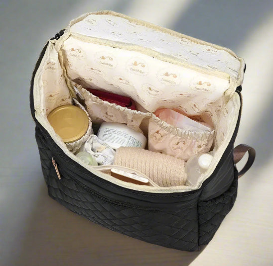 Un sac à langer noir | Baby Diaper est ouvert pour révéler un contenu organisé - des couches, un pot avec un couvercle doré, un bandage, des biberons et d'autres articles de soins pour bébé - soigneusement rangés dans des compartiments étiquetés.
