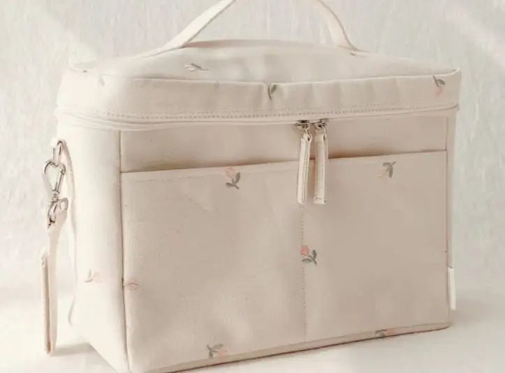 Le Sac à Langer Bébé est un sac en tissu rectangulaire de couleur crème avec une fermeture éclair, de petits imprimés floraux, une poche extérieure, une poignée supérieure et un fermoir latéral en métal, offrant une fonctionnalité élégante aux parents en déplacement.