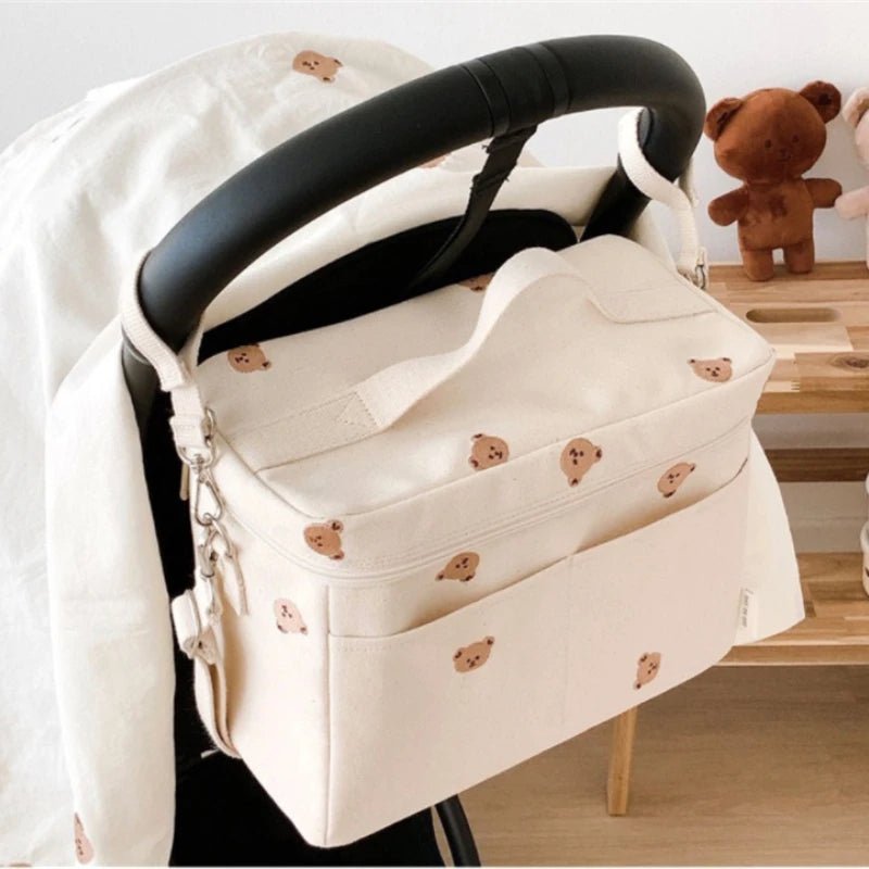Un Sac à Langer Bébé couleur crème, orné de petits visages d'ours, est suspendu à la poignée d'une poussette, alliant praticité et style. Derrière, un ours en peluche repose sur une étagère en bois.