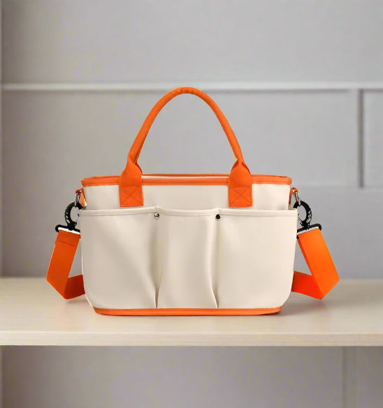 Le Sac à Langer | Bébé est un sac à langer rectangulaire beige avec poignées, bretelles et bordures orange. Il est doté de deux poches avant pour les accessoires de bébé et d'une bandoulière amovible, le tout sur fond blanc uni.
