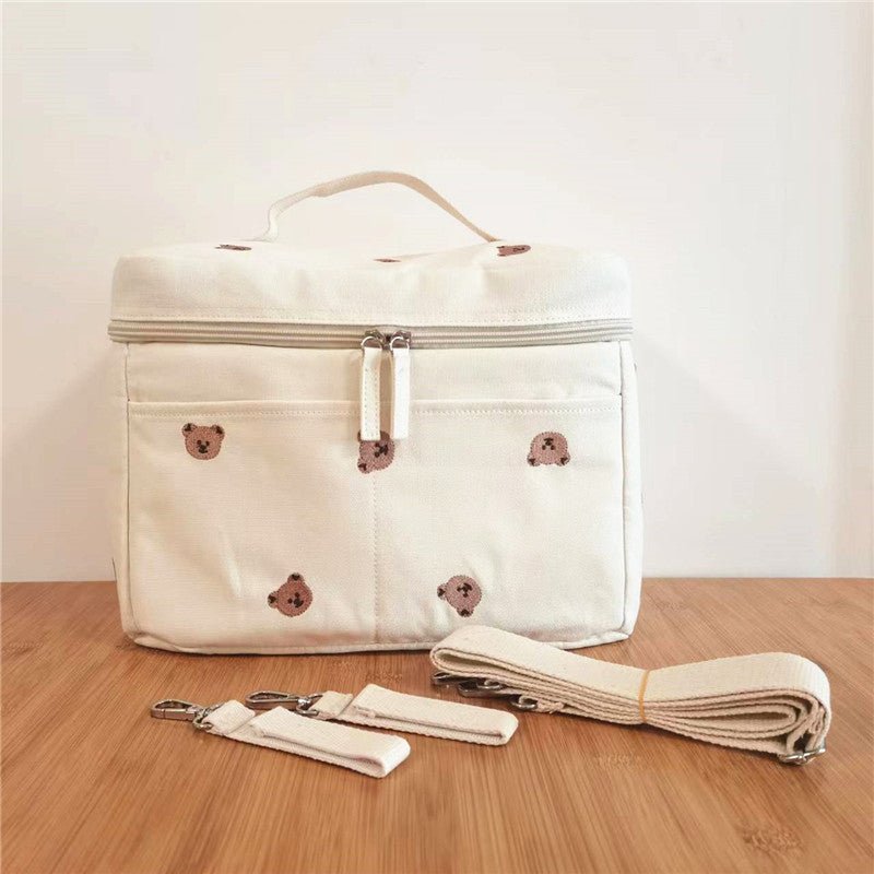 Le Sac à Langer Bébé de couleur crème, doté de petits visages d'ours bruns, d'une poignée et d'une fermeture éclair, repose sur une surface en bois avec deux sangles amovibles et une longue sangle assortie pour une fonctionnalité polyvalente.