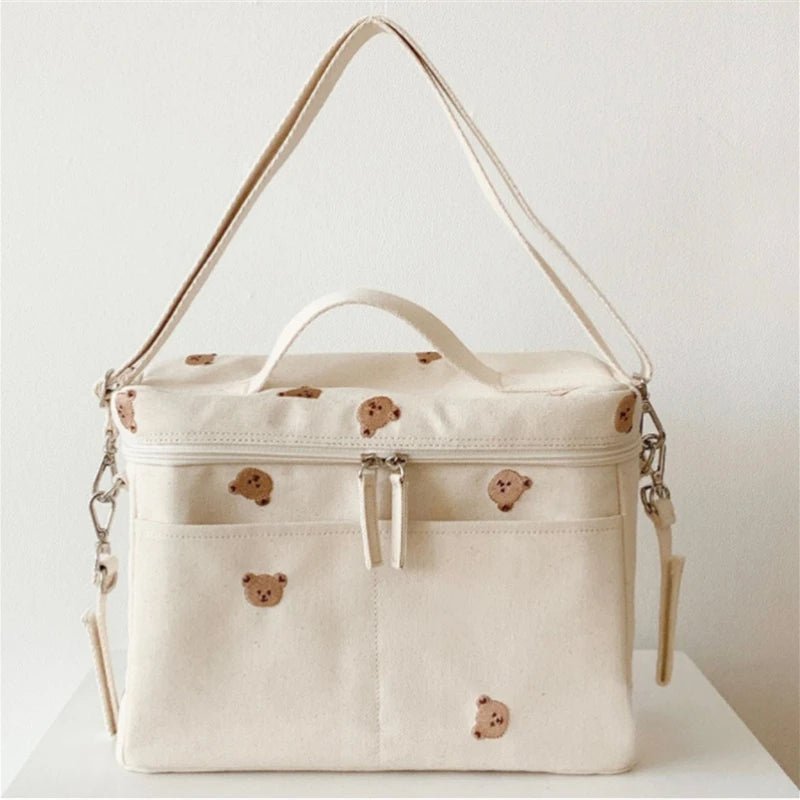Le Sac à Langer Bébé est un sac à langer en toile crème avec deux poignées, une fermeture éclair et d'adorables têtes d'ours bruns. Il est doté de deux poches avant, de détails argentés et d'une bandoulière amovible pour plus de style et de fonctionnalité.