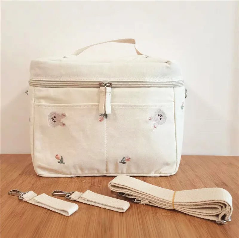 Le Sac à Langer Bébé est un sac à langer rectangulaire de couleur crème avec fermeture éclair, motifs subtils d'ours et de plantes, poignée supérieure et deux sangles amovibles, alliant style et fonctionnalité sur une surface en bois.