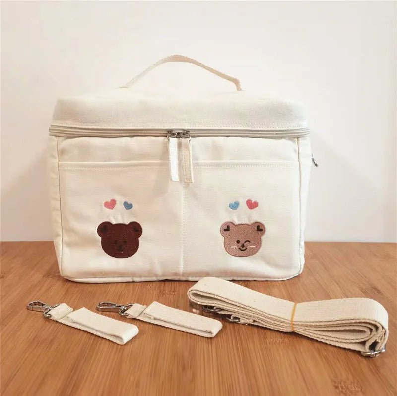 Le Sac à Langer Bébé crème comporte deux poches avant avec des visages d'ours et des cœurs brodés, ainsi que trois sangles amovibles pour plus de style et de fonctionnalité, présentées sur une surface en bois sur un fond uni.