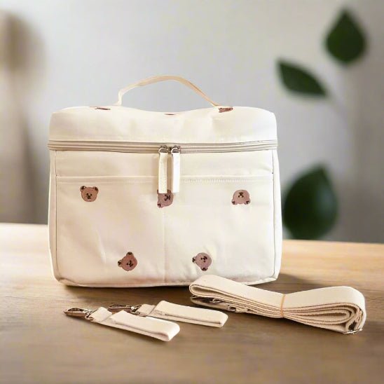 Le Sac a Langer Bebe - TeddyBear Organizer, un sac à langer rectangulaire de couleur crème avec poignée et fermeture éclair, est orné de petits visages d'ours. Deux sangles et pinces assorties sont placées devant sur une surface en bois, avec une plante floue en arrière-plan.