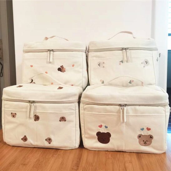Quatre sacs isothermes beiges Sac à Langer Bébé, ornés de motifs d'ours, de fermetures à glissière et d'accents de cœur colorés, sont disposés sur un plancher en bois. Chaque sac comprend une poignée supérieure pour un transport facile, alliant fonctionnalité et style sans effort.