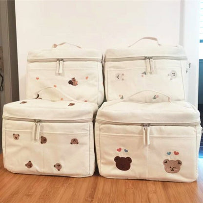 Quatre sacs isothermes beiges Sac à Langer Bébé, ornés de motifs d'ours, de fermetures à glissière et d'accents de cœur colorés, sont disposés sur un plancher en bois. Chaque sac comprend une poignée supérieure pour un transport facile, alliant fonctionnalité et style sans effort.