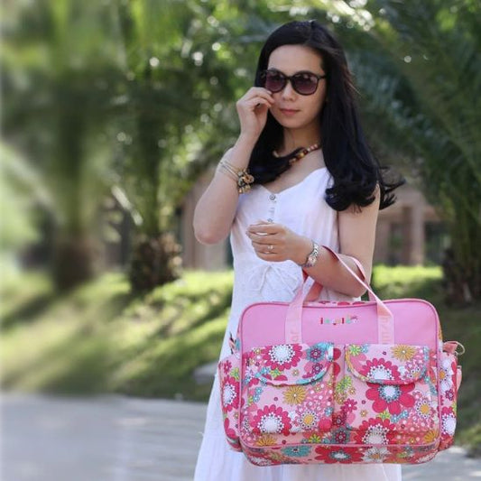 Une femme vêtue d'une robe blanche sans manches et de lunettes de soleil se tient à l'extérieur avec de la verdure derrière elle, tenant le Sac à langer bébé - Girly, un sac à langer coloré à motifs floraux, idéal pour les mamans modernes grâce à sa conception imperméable.
