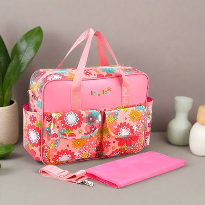 Le Sac à langer bébé - Girly présente un motif floral rose, plusieurs poches, des poignées roses, une bandoulière amovible et comprend un matelas à langer assorti, idéal pour les mamans modernes qui veulent à la fois style et praticité.