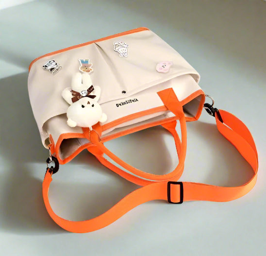 Le Sac à langer | Bébé beige, doté de bretelles orange et de pins en peluche et émail, est idéal pour les parents actifs. Présenté sur fond blanc.